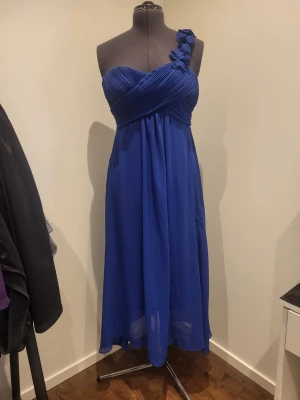 Blå långklänning med oneshoulder - Elegant blå långklänning med oneshoulder och dekorativa volangdetaljer på axeln. Klänningen har draperad överdel och ett luftigt, svepande tyg som ger en snygg siluett. Perfekt för speciella tillfällen när du vill sticka ut. Stl 36-38, bröst ca 97, längd ca 114 från armhåla. Strechigt tyg