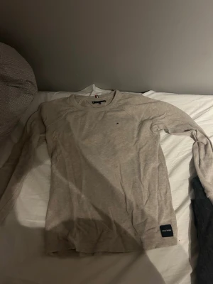 Beige långärmad tröja Tommy Hilfiger - Snygg beige långärmad tröja från Tommy Hilfiger med diskret logga på bröstet och en liten patch vid nederkanten. Tillverkad i mjukt material som känns skönt mot huden. Perfekt till jeans eller chinos för en clean look.