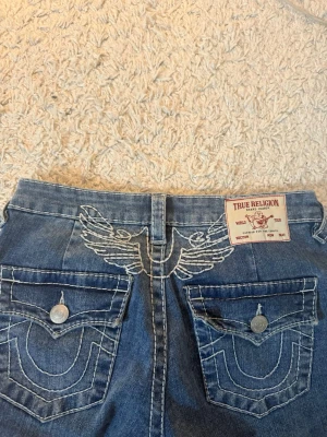 True Religion bootcut jeans med vingar - Säljer ett par blå bootcut jeans från True Religion med ikoniska vita kontrastsömmar och broderad logga på bakfickorna. Jeansen har låg midja, ficklock med knappar och en snygg vinge-detalj bak. Klassisk denim-look som passar till allt. Storlek 26. 