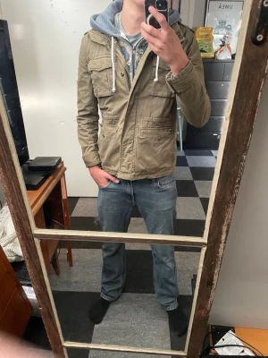 Olivgrön vindjacka från Superdry - Säljer en olivgrön vindjacka från Superdry med fyra stora fickor framtill och huva. Jackan har dragkedja och knappar en knapp är borta , samt en cool militärinspirerad look. Materialet är slitstarkt och jackan är perfekt för blåsiga dagar.