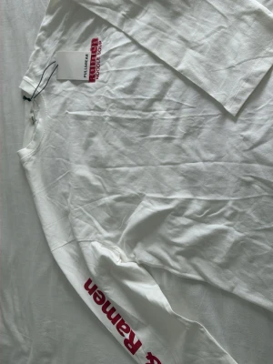 Vit långärmad Ramen t-shirt Pull&Bear - Vit långärmad t-shirt från Pull&Bear med röd text 'Ramen Noodle Soup' på bröstet och 'Ramen' tryckt längs ena ärmen. Tillverkad i mjuk bomull och har en klassisk rund hals. Perfekt för dig som gillar streetwear och snygga prints.