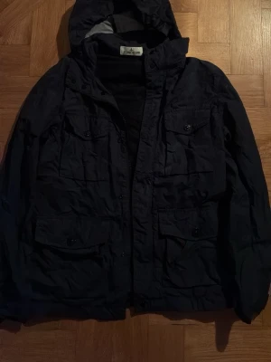 Stone Island Svart vindjacka med huva - Snygg stone island svart vindjacka med huva och flera praktiska fickor framtill. Jackan har dragkedja och knappar, samt justerbar huva. Perfekt för blåsiga dagar och enkel att matcha med olika outfits.