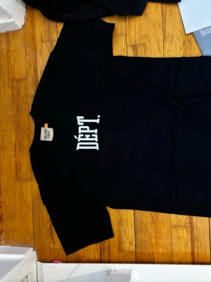 Svart t-shirt från Gallery Dept - Svart t-shirt från Gallery Dept med stort vitt tryck 'DÉPT.' på både fram- och baksidan. Klassisk passform och rund hals. Perfekt för dig som gillar streetwear och vill ha en enkel men snygg look.