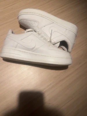 Helvita Air Force 1 sneakers - Säljer ett par klassiska helvita Air Force 1 sneakers i läder. Skorna har den ikoniska swoosh-loggan på sidan, perforerad tå och tjock sula med AIR-detalj. Perfekta för dig som gillar stilrena och tidlösa sneakers. Priset kan diskuteras 