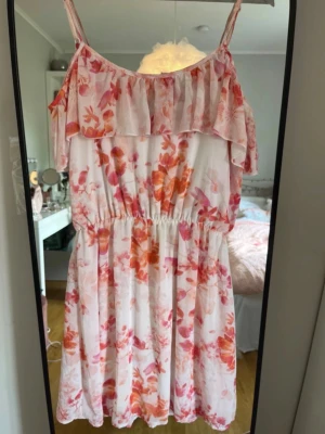 Blommig sommarklänning med volang - Söt kortklänning med blommigt mönster i rosa och orange toner. Klänningen har smala axelband, volang upptill och resår i midjan för en bekväm passform. Perfekt för varma dagar och somriga tillfällen.