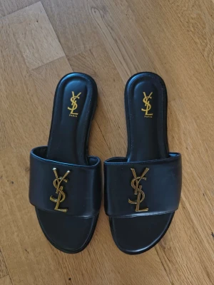 Svarta YSL sandaler i läder - Snygga svarta sandaler från Yves Saint Laurent med bred rem i äkta läder och ikonisk YSL-logga i guld framtill. Slip-in modell med öppen tå och platt sula, perfekt för en trendig och lyxig look. Klassisk design som passar till många outfits.