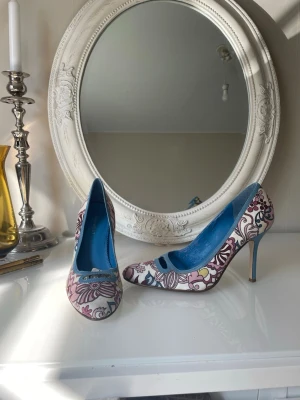 Damskor Nine West  - Unika pumps från Nine West med färgglatt blommönster i rosa, lila och vitt. Skorna har spetsig tå, smal klack och blå insida samt dekorativ blå kant framtill. Perfekta för dig som vill sticka ut med din stil.