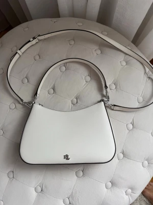 Vit axelremsväska från Ralph Lauren - Ny stilren vit handväska i slätt konstläder med silverfärgade detaljer och RL-logga framtill. Väskan har både ett kort handtag och en justerbar axelrem, vilket gör den flexibel att bära. Enkel, modern design med svarta kanter för extra kontrast. Endast använd 2 gånger, inte en skråma på den. Nypris var 3000kr. Dustbag ingår.