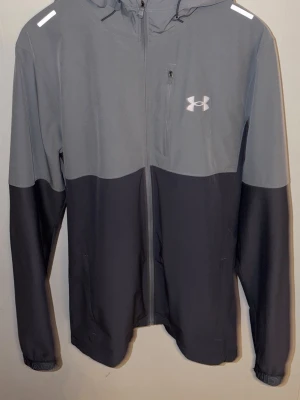 Grå vindjacka från Under Armour - Snygg tvåfärgad vindjacka från Under Armour i ljus- och mörkgrått. Jackan har huva, hel dragkedja, bröstficka med dragkedja och reflekterande detaljer på axlarna. Perfekt för blåsiga dagar och sportiga outfits. 