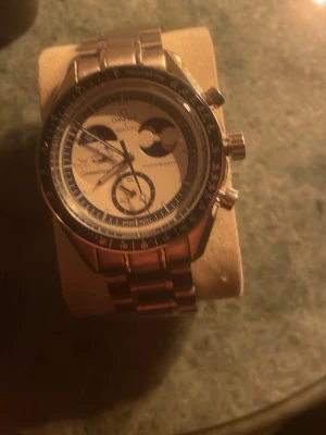Omega Speedmaster Moonwatch klocka - Säljer en Omega Speedmaster Moonwatch med blå och vit urtavla, detaljerad med månfas och kronograf. Klockan har robust boett i silverfärgat stål och finns med både länkarmband i metall och blått textilarmband. Klassisk och ikonisk design för dig som gillar stilrena accessoarer.