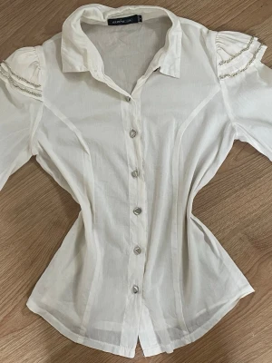 Vit blus med puffärm och pärldetaljer - Stilren och feminin blus från Susie Young i en mjuk krämvit nyans. Den har en figursydd passform som framhäver midjan och dekorativa silverkedjor på axlarna som ger ett exklusivt uttryck. Blusen stängs med triangelformade metallknappar framtill och har en klassisk krage som gör den perfekt både till vardag och fest. Passar utmärkt med kostymbyxor eller kjol för en elegant, old-money inspirerad look. Tyget är lätt och har en subtil glans. Uppskatar storlek XS. Jättefin material. Känns fint och ri