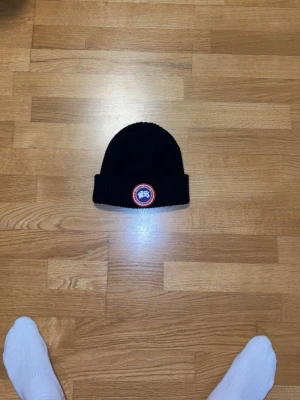  Canada Goose - Säljer en svart ribbstickad mössa från Canada Goose med uppvikt kant och klassisk rund logotyp framtill. Perfekt för kalla dagar och enkel att matcha med olika outfits. Tidlös design som funkar året runt.