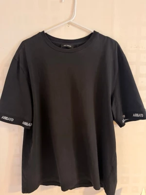 Svart t-shirt från Axel Arigato - Svart t-shirt från Axel Arigato med rund hals och korta ärmar. På ärmsluten finns vit text med märkesnamnet 'ARIGATO' broderat. Tillverkad i mjuk bomull för en skön och avslappnad stil.