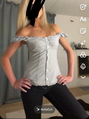 Randig offshoulder blus med knappar - Snygg offshoulder blus med smala blå och vita ränder. Toppen har korta puffärmar, knappar framtill och volangkant vid ärmar och nederkant. Perfekt för en trendig och somrig look.