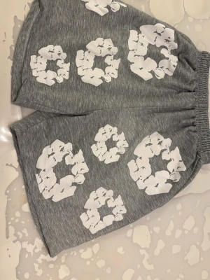 Grå shorts med vit grafisk print, den är blöt när jag tog bilden men den är mycket ljusare - Säljer ett par grå shorts med bred resår i midjan och stora vita grafiska tryck över hela plagget. Shortsen har en loose passform och är tillverkade i mjukt material som känns skönt mot huden. Perfekta för en avslappnad och trendig look.