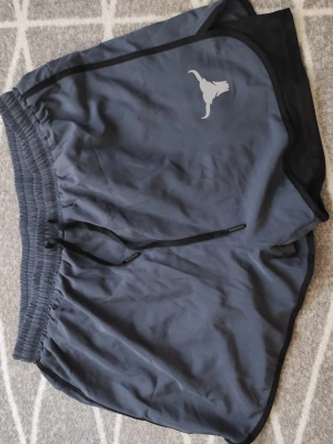 Mörkblå träningsshorts med snörning - Snygga mörkblå träningsshorts med elastisk midja och svart snörning. Shortsen har en vit tjur-logga på ena benet och svarta detaljer längs kanterna. Perfekta för gymmet eller löprundan. Små i storleken