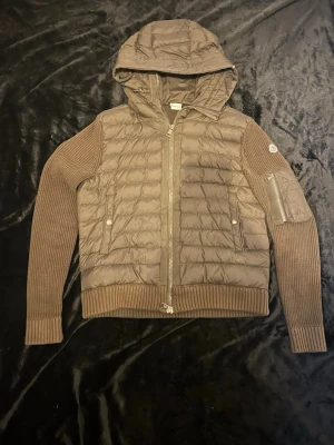 Moncler cardigan - Snygg olivgrön gardigan från Moncler med stickade ärmar och huva. Jackan har quiltad kropp, ribbade muddar och dragkedja framtill. Fickor med tryckknappar och en extra ficka på ärmen. Perfekt för kalla vinterdagar. Storlek:M
