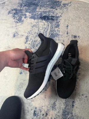 Svarta Adidas Ultraboost sneakers - Snygga svarta Adidas Ultraboost sneakers med vit sula och klassiska tre ränder på sidan. Ovandelen är i ventilerande mesh och skon har en bekväm Boost-mellansula. Perfekta för dig som gillar stilrena och sportiga sneakers.