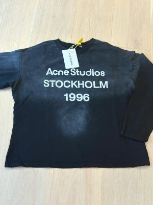 Svart långärmad t-shirt Acne Studios - Cool svart långärmad t-shirt från Acne Studios med vit text 'Acne Studios STOCKHOLM 1996' på bröstet. Klassisk rak passform och rund halsringning. Perfekt för dig som gillar streetwear och enkel stil.   Hör av dig vid minsta fundering😊