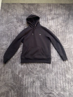 Svart hoodie från GANT med huva - Säljer en svart hoodie från GANT i storlek M. Hoodien har en klassisk känguruficka framtill, dragsko i huvan och en liten GANT-logga broderad på bröstet. Tillverkad i mjukt bomullsmaterial som är skönt mot huden. Perfekt för en avslappnad och stilren look.