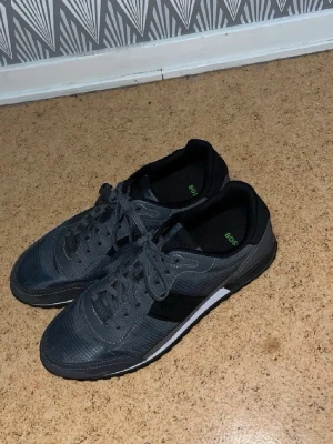 Mörkgrå sneakers från BOSS - Snygga mörkgrå sneakers från BOSS med svarta detaljer och vita inslag på sulan. Skorna har klassisk låg profil, snörning och en stilren design. Materialet ser ut att vara en mix av textil och syntet, perfekt för en modern och avslappnad look.