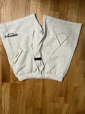 Grå mjukis shorts från Essentials - Snygga ljusgrå mjukisbyxor från Essentials med svart logga på vänster lår. Byxorna har resår och dragsko i midjan samt muddar vid bensluten. Perfekta för en avslappnad och trendig look.