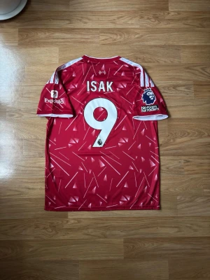 Liverpool hemma matchtröja Isak #9 | storlek: M - Säljer en röd Liverpool matchtröja med vita detaljer och grafiskt mönster. Tröjan har trycket 'Isak' och nummer 9 på ryggen, Premier League-märke på ärmen samt sponsorloggor. Perfekt för fotbollsfans som vill sticka ut på läktaren eller planen.
