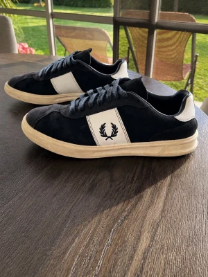 Svarta Fred Perry sneakers i mocka - Snygga svarta sneakers från Fred Perry med vit panel och logga på sidan. Skorna har en klassisk låg siluett, mörkblå snören och beige sula. Ovandelen är i mocka och textil, vilket ger en schysst retrovibe.