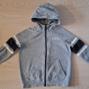 Grå Nike Air hoodie med svarta detaljer stl 147-158  - Snygg grå Nike Air hoodie med dragkedja, huva och svarta partier med vita ränder på ärmarna. Broderad Nike Air-logga på bröstet och två fickor framtill. Perfekt för chill dagar eller när du vill ha en sportig look.