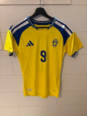 Sverige matchtröja Isak #9 Adidas storlek S - Gul och blå Sverige matchtröja  med nummer 9 och namnet Isak på ryggen. Tröjan har korta ärmar, blå detaljer på axlarna och klassiska Adidas-ränder. Perfekt för dig som vill stötta landslaget på match eller träning.