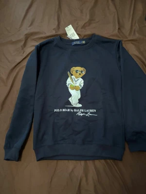 Svart sweatshirt Polo Bear Ralph Lauren - Cool svart sweatshirt från Ralph Lauren med Polo Bear-tryck på bröstet. Tröjan har rund halsringning och ribbade muddar vid ärmslut och nederkant. Perfekt för en avslappnad och trendig stil.