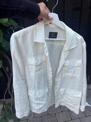 Vit Linné overshirt från Less Worries - Stilren vit overshirt från Less Worries i ett Linné material. Skjortan har fyra fickor framtill med lock och knappar, klassisk krage och långa ärmar. Perfekt för lager på lager och en avslappnad look. Aldrig använts men lapp e borttagen! Nypris 1500 kr.