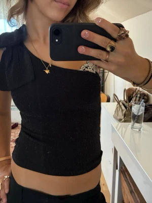 Svart oneshoulder topp med rosett - Säljer en svart oneshoulder topp med stor rosettdetalj på axeln. Toppen är glittrig och har en tight passform som framhäver figuren. Perfekt för dig som vill sticka ut lite extra med en unik detalj på axeln. Den är storlek 152 men passar mig som har s/xs