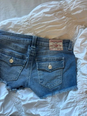 Blå jeansshorts från True Religion - Säljer ett par blå jeansshorts från True Religion med fransig kant och klassiska bakfickor med lock och silverknappar. Shortsen har normal passform och bälteshällor, perfekta för sommaren. Snyggt slitna detaljer och ikonisk logga bak.
