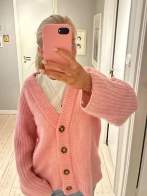 Rosa stickad kofta  - Mysig rosa stickad kofta med stora bruna knappar och djup v-ringning. Koftan har en avslappnad passform och breda ärmar, perfekt att slänga på sig över en t-shirt. Ribbstickade muddar och mjukt material ger en skön känsla.