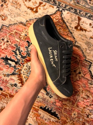 Svarta Saint Laurent sneakers - Svarta sneakers från Saint Laurent med handskriven logga på sidan. Skorna har en klassisk låg profil, svarta snören och kontrasterande beige sula. Tillverkade i canvas med detaljer i läder. Perfekta för en stilren och trendig look.