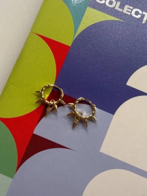 Guldiga hoops med nitar - Säljer ett par små guldiga hoop-örhängen med coola spetsiga nitar runt hela ringen. Perfekta för dig som vill ha en edgy och trendig look. Materialet ser ut att vara metall med guldfinish och designen är riktigt modern.