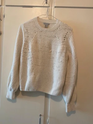 Vit stickad tröja från H&M - Säljer en krämvit stickad tröja från H&M med rund halsringning och ribbade muddar. Tröjan har ett diskret hålmönster vid axlarna och är tillverkad i ett mjukt, tjockt material som passar perfekt för kyligare dagar.
