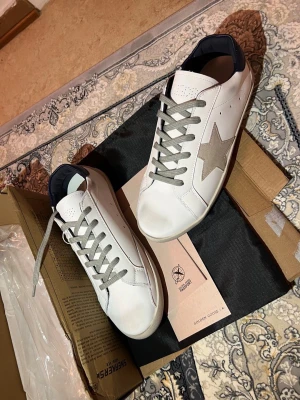 Vita sneakers med stjärndetalj - Snygga vita sneakers från Golden Goose med grå stjärna på sidan, grå skosnören och mörkblå hälflik. Skorna har en klassisk låg profil och är tillverkade i läder med mockadetaljer. Perfekta för en trendig och avslappnad stil.