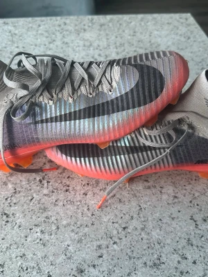 Nike Mercurial CR7 fotbollsskor - Nike Mercurial CR7 fotbollsskor i grått med svart swoosh och CR-detalj på sidan. Skorna har ribbad ovandel, snörning och en orange/röd sula med fasta dobbar. Tillverkade i syntetmaterial för bästa grepp och känsla på planen.