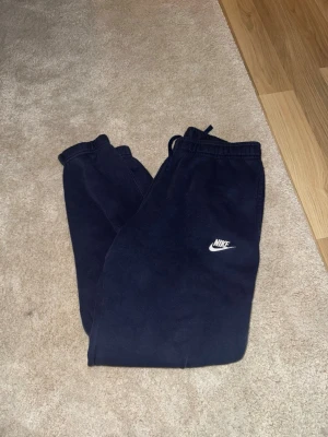 Mjukisbyxor - This is a pair of navy blue Nike sweatpants with a drawstring waistband and the Nike logo on the left thigh. The pants are laid flat on a carpeted floor, with one leg crossed over the other. The material appears to be soft and comfortable, suitable for casual wear or athletic activities.