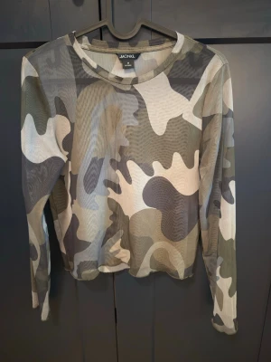 Camouflage mesh topp från Monki - Säljer en tunn och transparent långärmad topp från Monki med camouflage-mönster i gröna, beige och grå toner. Toppen är figurnära och har rund hals. Perfekt att styla lager på lager för en cool look.