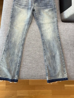 Blå flared jeans från HESNTLS - Säljer ett par blå jeans från HESNTLS med slitna detaljer och ljusa tvättade partier. Jeansen har raka ben, råa kanter vid benslut och coola färgstänk för en trendig look. Perfekta för dig som gillar streetstyle och vill sticka ut.