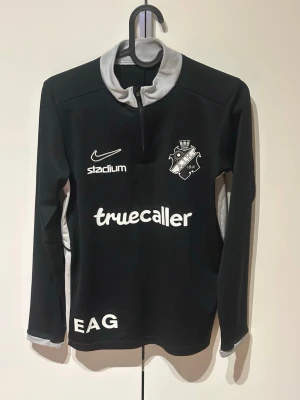 Svart AIK träningströja Nike - Svart långärmad träningströja från Nike med AIK:s klubbmärke på bröstet. Tröjan har halv dragkedja, grå krage och detaljer samt vita loggor och tryck. 