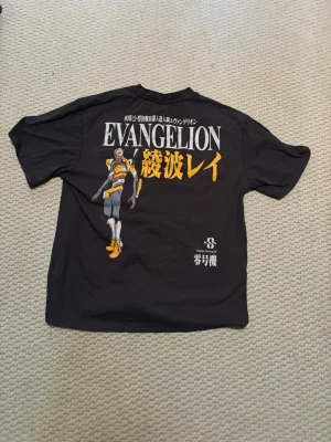 Svart Evangelion t-shirt med tryck - Cool svart t-shirt med stort Evangelion-tryck på ryggen och text på både fram- och baksida. Motivet visar en karaktär från serien och text i vitt och gult. Perfekt för animefans som vill sticka ut med en unik tee.
