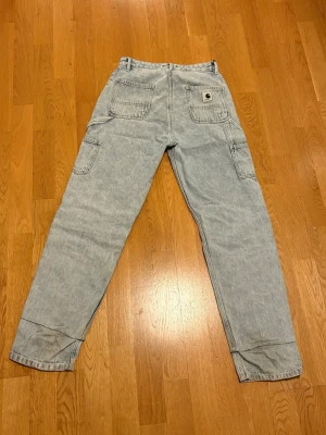 Ljusblå Carhartt workwear jeans - Säljer ett par ljusblå workwear jeans från Carhartt med raka ben och flera praktiska fickor. Jeansen har en avslappnad passform och är tillverkade i slitstark denim. Perfekta för en streetwear-look eller till vardags.