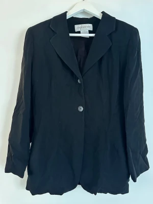 Svart kavaj med två knappar - Stilren svart kavaj från Jones New York med klassisk krage och två knappar framtill. Lång ärm och rak passform. Perfekt för en clean och enkel look. Materialet ser ut att vara lätt och mjukt, vilket ger en avslappnad känsla. USA STORLEK 10, motsvarar runt storlek M.