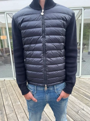 Svart dunväst från Moncler - Snygg svart dunväst från Moncler med quiltad front och mjuka stickade ärmar. Västen har dragkedja framtill, två sidofickor med dragkedja och Moncler-logga på ärmen. Perfekt för lager-på-lager och streetstyle.