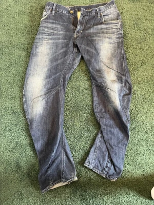 Blå jeans från G-Star med tvätt - Säljer ett par blå jeans från G-Star med snygga slitningar och tvättade detaljer. Jeansen har raka ben, fem fickor och coola sömmar längs benen. Materialet är klassisk denim och passformen är avslappnad. Perfekta för en trendig och avslappnad stil. W:32