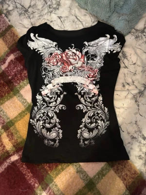 Svart t-shirt med drak- och rosprint - Cool svart t-shirt med korta ärmar och detaljerat tryck framtill med vita drakar, silverfärgade mönster och röda rosor. Slimmad passform som sitter snyggt. Perfekt för dig som gillar edgy och unika plagg. Kragen är avklippt själv 